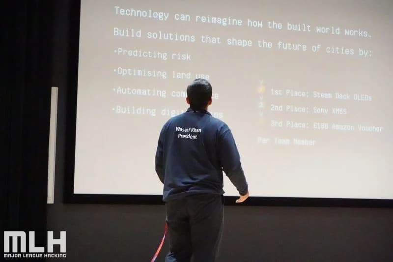 A HackLondon organiser presents challenge results to participants in an auditorium.